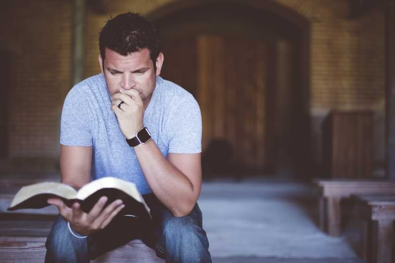 A sitting man reads an open Bible, holding it in his right hand while his left hand sits on his chin in a thoughtful pose. © By Ben White/Wirestock/stock.adobe.com