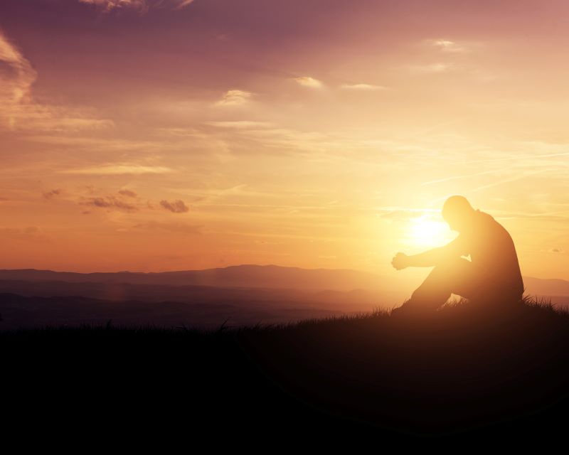 A man sits on a small hillside with his hands folded in prayer at sunset. © By Kevin Carden/stock.adobe.com